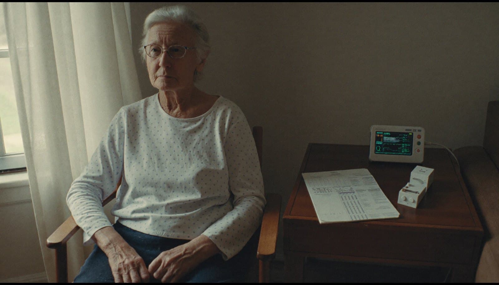 An older adult recently home from the hospital, a connected home monitoring device and discharge paperwork on the side table beside them